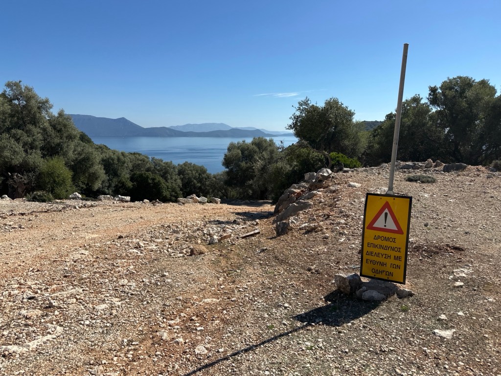 A sign warning of a dangerous road for vehicles, set against a backdrop of a scenic view of the sea and mountains on Meganisi island.