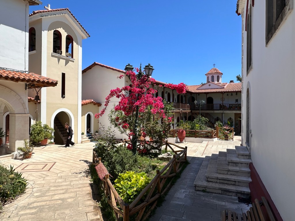A view of a monastery courtyard in Lefkada, featuring white buildings with terracotta roofs, vibrant pink bougainvillea flowers, and green plants. A person walks through the courtyard under a clear blue sky.