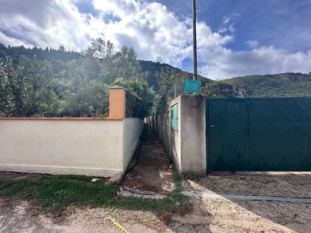 A narrow path flanked by walls leading to a green gate, with scenic hills in the background and blue skies.