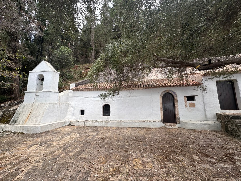 A small white church with a bell tower surrounded by trees, located on a stone path. The building has a traditional Mediterranean architectural style.