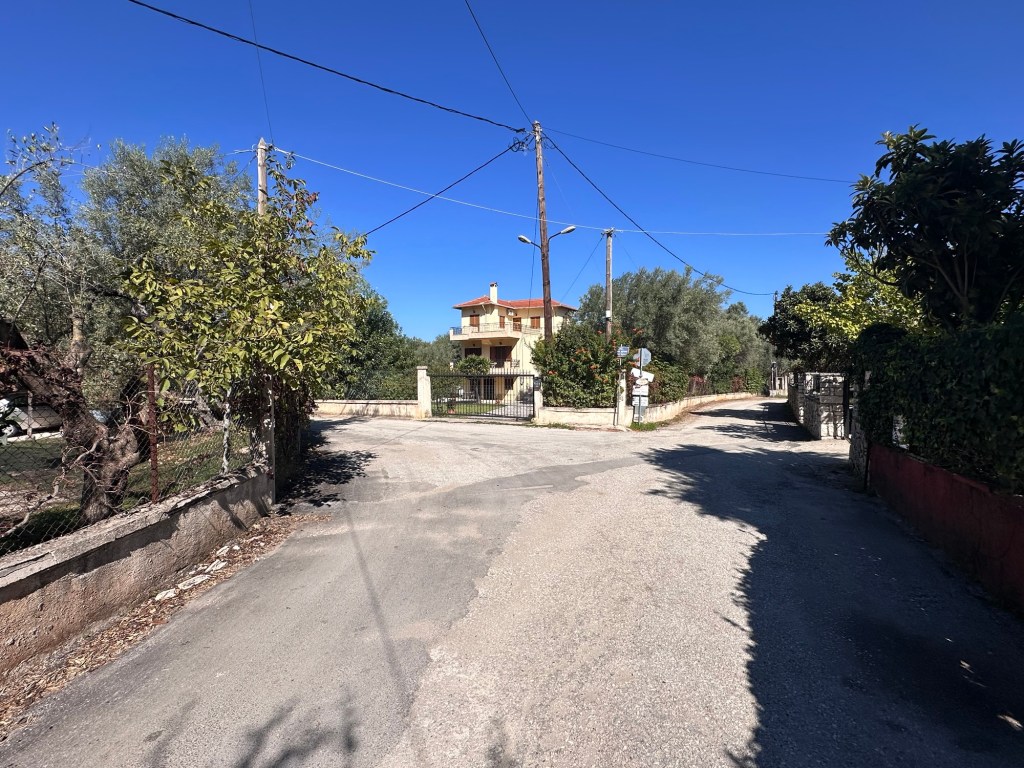 A rural intersection in Lefkada, Greece, featuring a dirt road leading to a residential building surrounded by trees and vegetation under a clear blue sky.